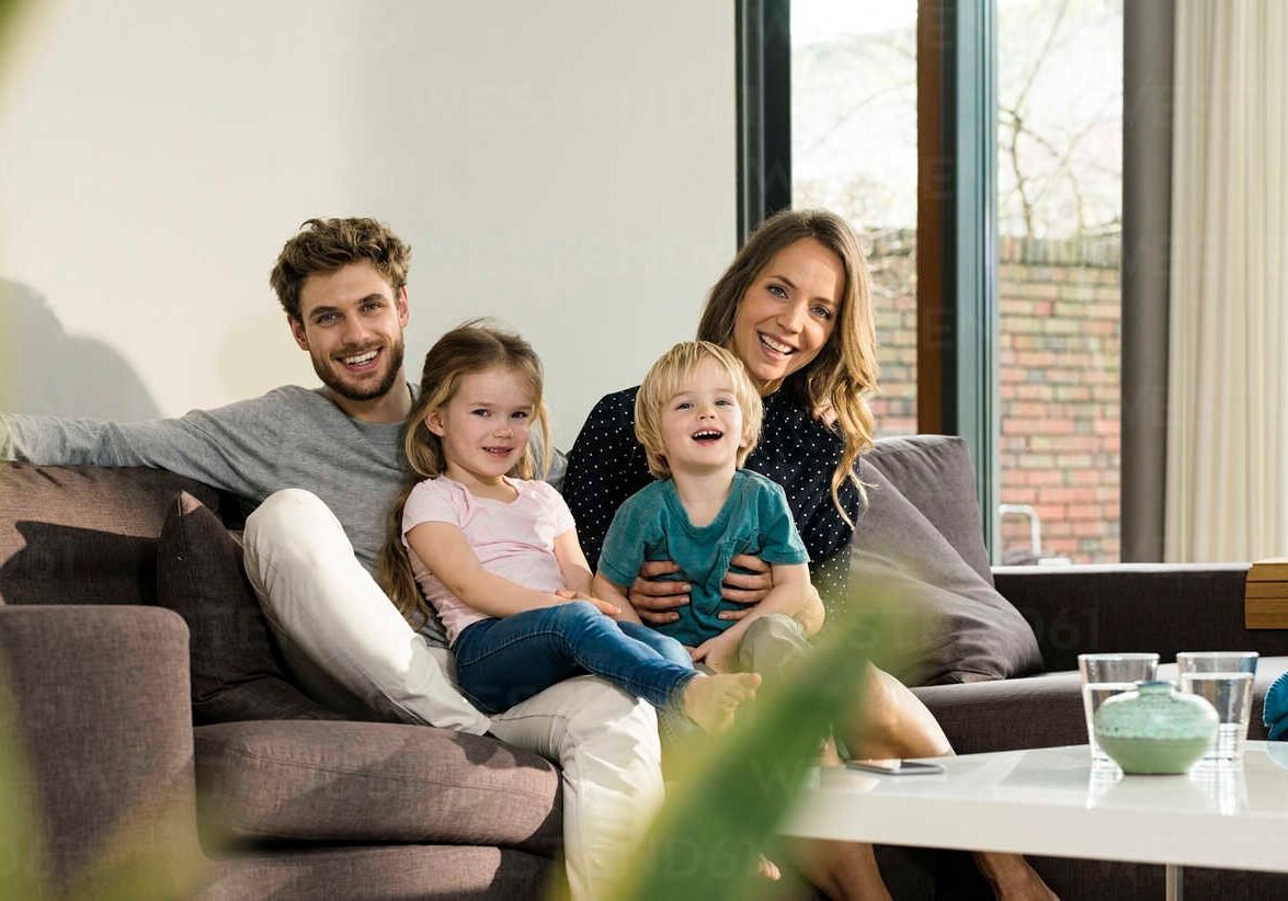 portrait-of-happy-family-sitting-on-sofa-at-home-SBOF01301 Germany, smiling family of mother, father, toddler son and daughter having fun and sitting on sofa in modern design living room with large garden windows